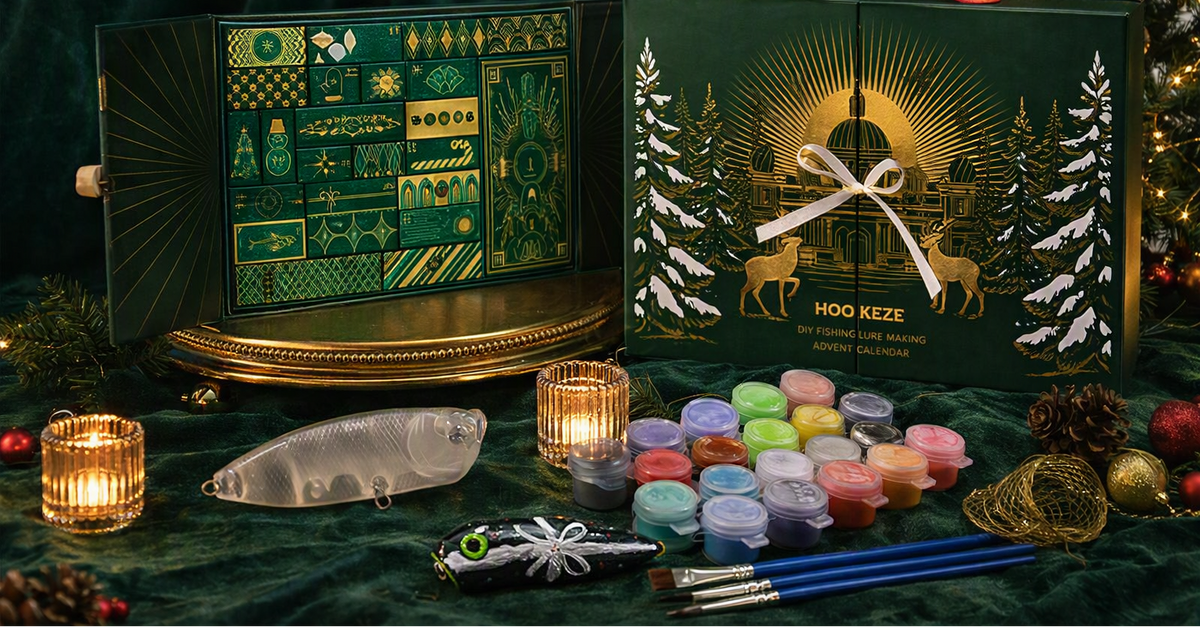 A festive Hogg Keze Christmas Advent Calendar set displayed on a dark green velvet surface with holiday decorations. The set includes an open calendar showing green and gold interior pages, a closed dark green gift box with gold artwork and a red ribbon handle, multiple small paint pots, a fish-shaped object, and paintbrushes.
